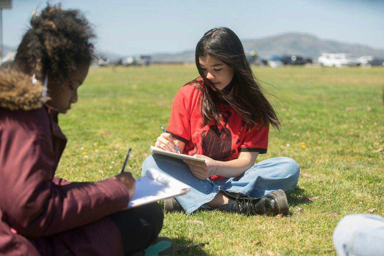 two ati museum mile students sitting on grass writing
